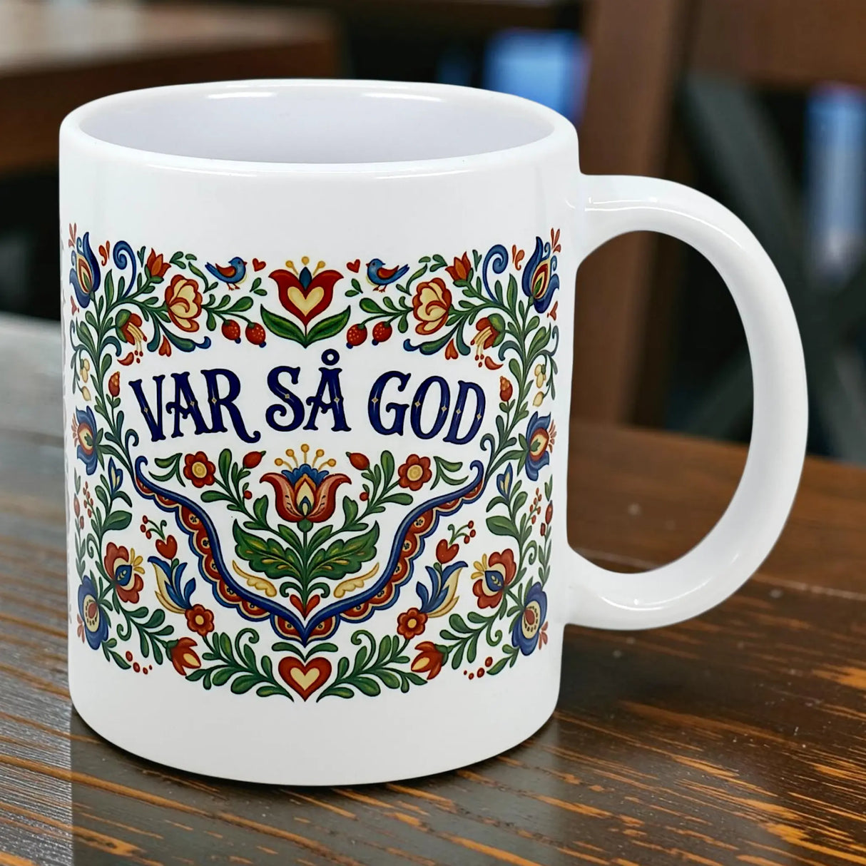 The Mug: Var Sa God Swedish Kurbits Folk Art Ceramic Mug (11 oz) by Nordic Wholesalers features colorful floral patterns and "VAR SÅ GOD" in the center, inspired by Scandinavian design, displayed on a wooden table.
