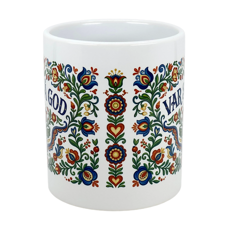 The Mug: Var Sa God Swedish Kurbits Folk Art Ceramic Mug (11 oz) by Swedish Gift Store is a white mug featuring colorful, symmetrical floral patterns and blue "GOD VAR" text, inspired by traditional Scandinavian folk art.