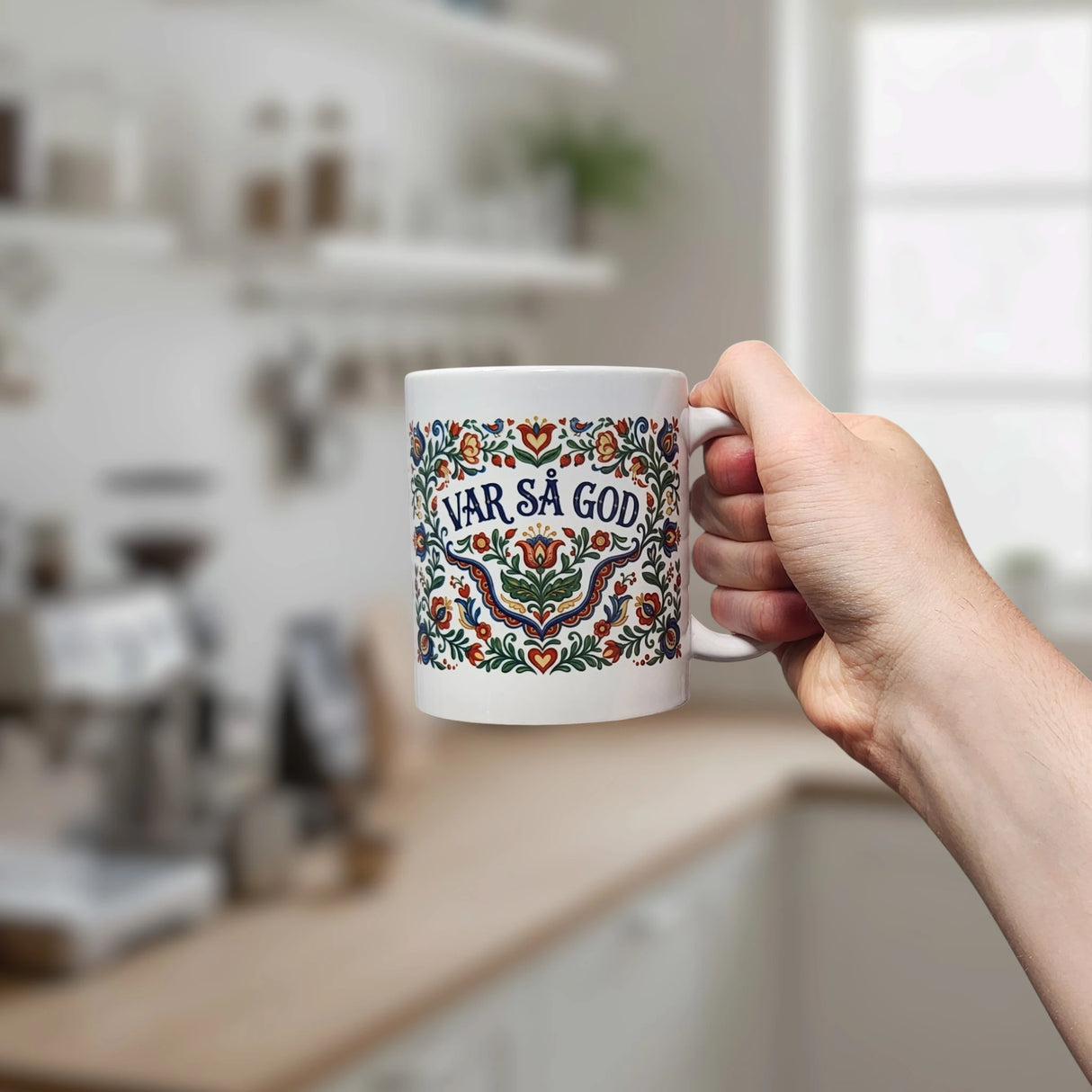 A hand holds the Nordic Wholesalers Mug: Var Sa God Swedish Kurbits Folk Art Ceramic Mug (11 oz), featuring colorful floral designs and the phrase VAR SÅ GOD, in a bright modern kitchen with blurred shelves and a window in the background.