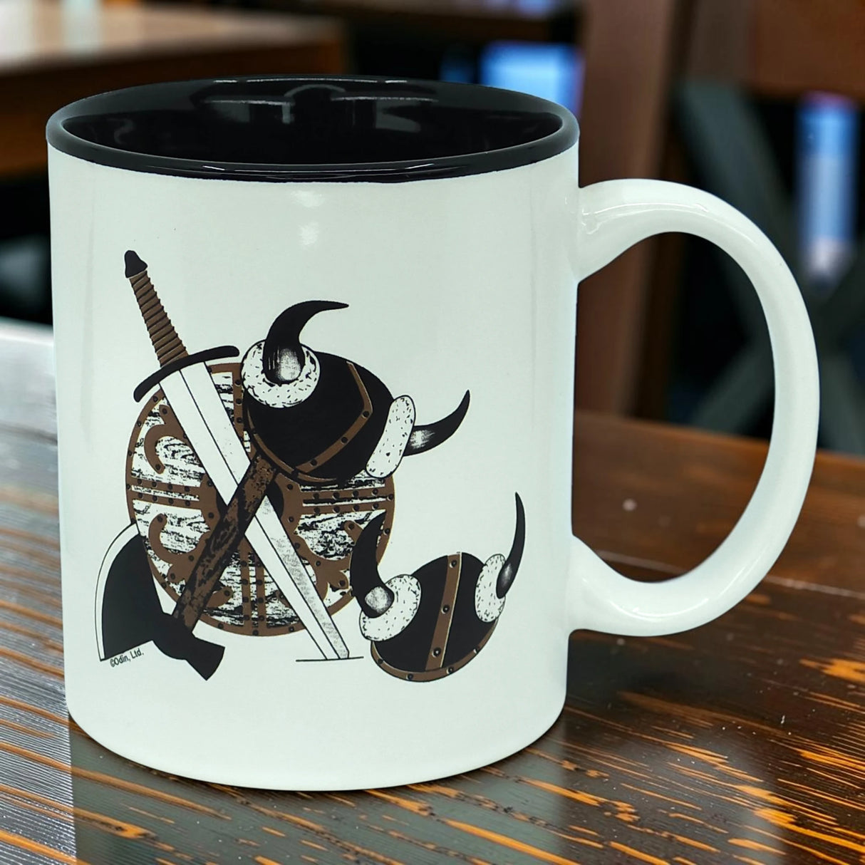 Introducing the Mug: Viking Coat of Arms | Black Interior | 11oz Ceramic Mug by Odin Limited—a white ceramic mug with a bold black interior, showcasing an illustration of Viking helmets, sword, and shield arranged together on one side. This striking design pays tribute to Norse warrior heritage. The Odin Limited Viking mug is pictured resting on a wooden surface with blurred chairs in the background.