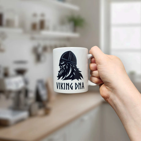 A hand holds the 11 oz Mug: Viking DNA White Ceramic Mug with Historically Accurate Helmet by Nordic Wholesale, featuring a black illustration of a Viking helmet and beard alongside the text "VIKING DNA," in a blurred kitchen setting.