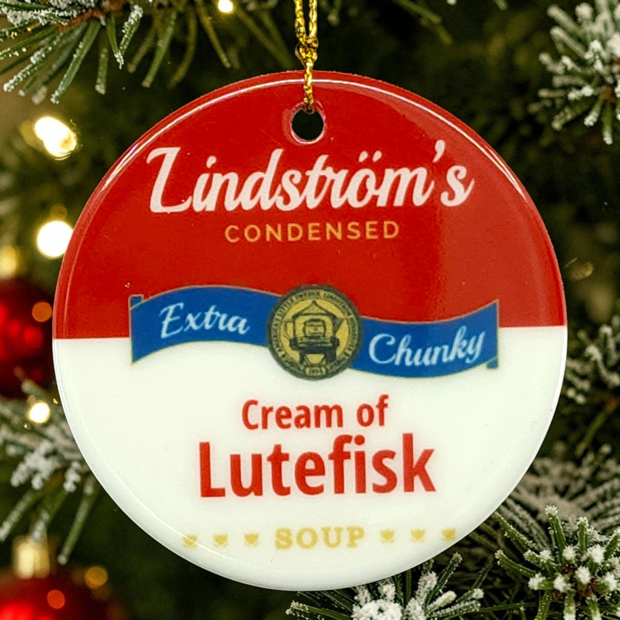 A round Nordic ornament designed like a soup can label, with the text “Lindströms Cream of Lutefisk Soup,” hanging from a branch of a decorated Christmas tree.