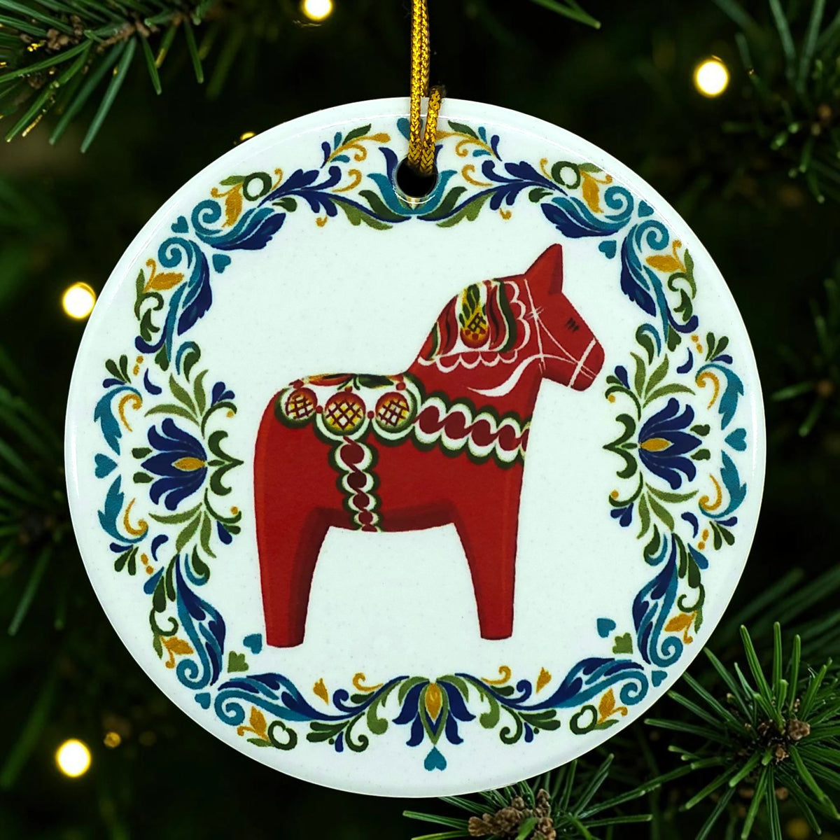 Round white ceramic ornament featuring a Swedish Dala horse and colorful floral border, hanging by a gold string from a pine tree.