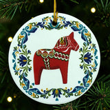 Round white ceramic ornament featuring a Swedish Dala horse and colorful floral border, hanging by a gold string from a pine tree.
