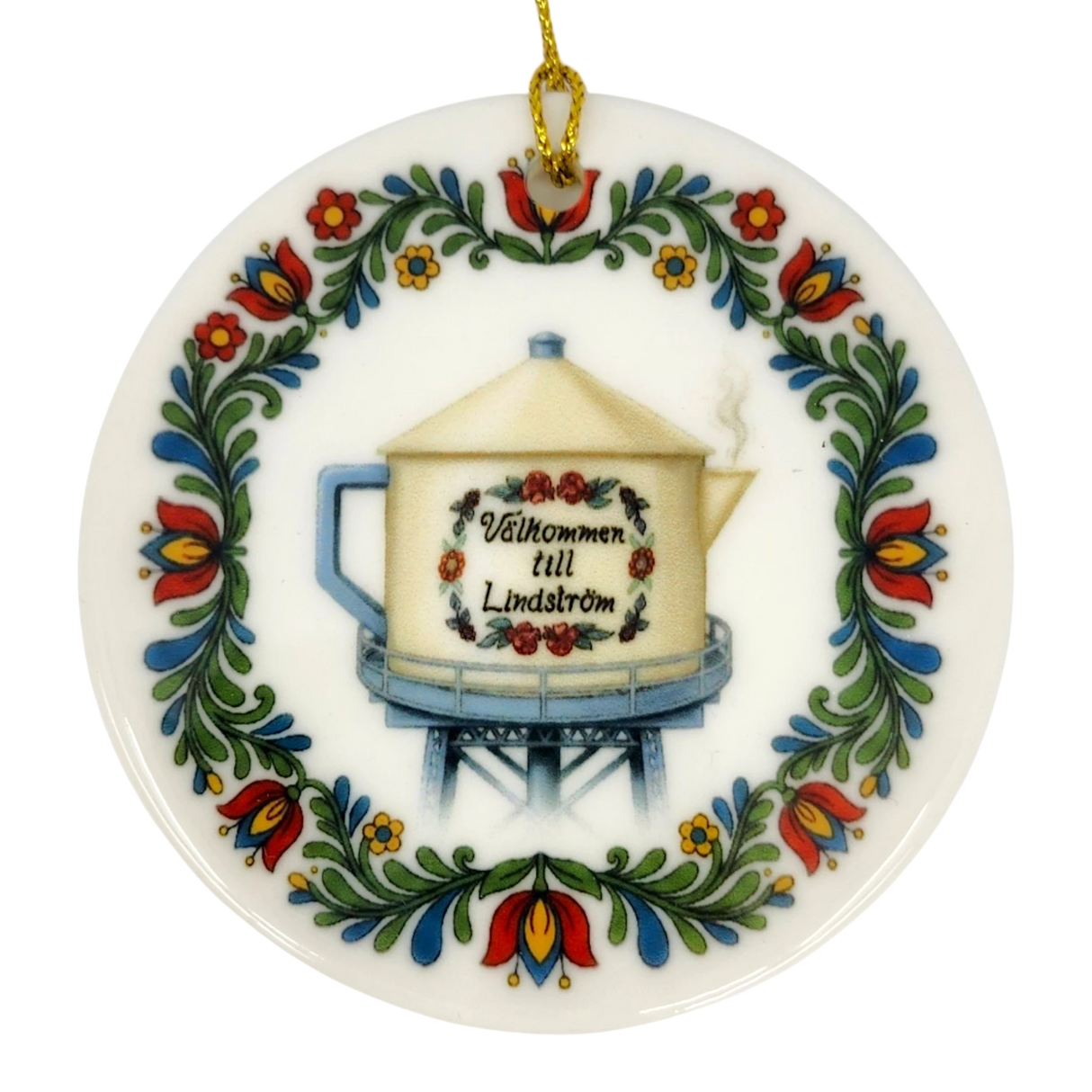 The Nordic Wholesale Ornament: Lindstrom Water Tower Floral Ceramic Ornament (2.875" Diameter) is a round Swedish ceramic ornament featuring a kurbits folk art floral border and the iconic Lindström water tower in the shape of a teapot, accented with the Swedish text “Välkommen till Lindström.” A gold string is attached at the top for hanging.