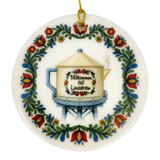 The Nordic Wholesale Ornament: Lindstrom Water Tower Floral Ceramic Ornament (2.875" Diameter) is a round Swedish ceramic ornament featuring a kurbits folk art floral border and the iconic Lindström water tower in the shape of a teapot, accented with the Swedish text “Välkommen till Lindström.” A gold string is attached at the top for hanging.