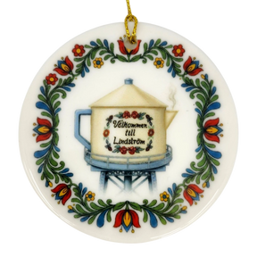 The Nordic Wholesale Ornament: Lindstrom Water Tower Floral Ceramic Ornament (2.875" Diameter) is a round Swedish ceramic ornament featuring a kurbits folk art floral border and the iconic Lindström water tower in the shape of a teapot, accented with the Swedish text “Välkommen till Lindström.” A gold string is attached at the top for hanging.