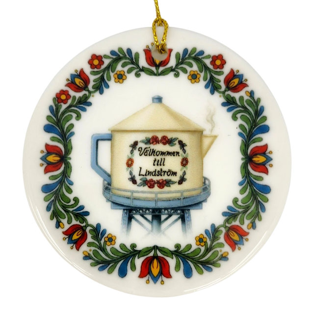 The Nordic Wholesale Ornament: Lindstrom Water Tower Floral Ceramic Ornament (2.875" Diameter) is a round Swedish ceramic ornament featuring a kurbits folk art floral border and the iconic Lindström water tower in the shape of a teapot, accented with the Swedish text “Välkommen till Lindström.” A gold string is attached at the top for hanging.