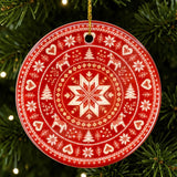 Red circular Nordic ornament featuring white snowflake, reindeer, and tree designs, hanging on a green Christmas tree with small white lights.
