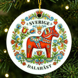 Round ornament with a red Dala horse decorated in colorful floral patterns and the words SVERIGE DALAHÄST, shown hanging on a Christmas tree branch.