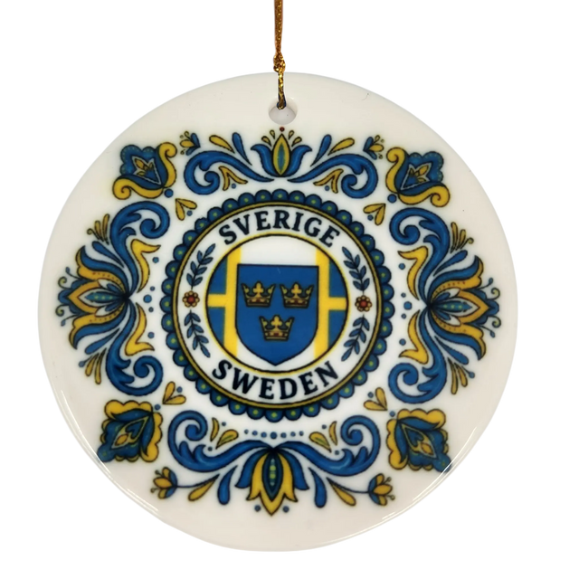The Ornament: Sverige Sweden Triple Crown Ceramic Ornament (2.875" Diameter) by Nordic Wholesale showcases blue and yellow floral patterns surrounding a central shield with three crowns and the words "Sverige Sweden." A gold string is attached at the top, making this Swedish ceramic ornament a charming piece of Nordic holiday decor.