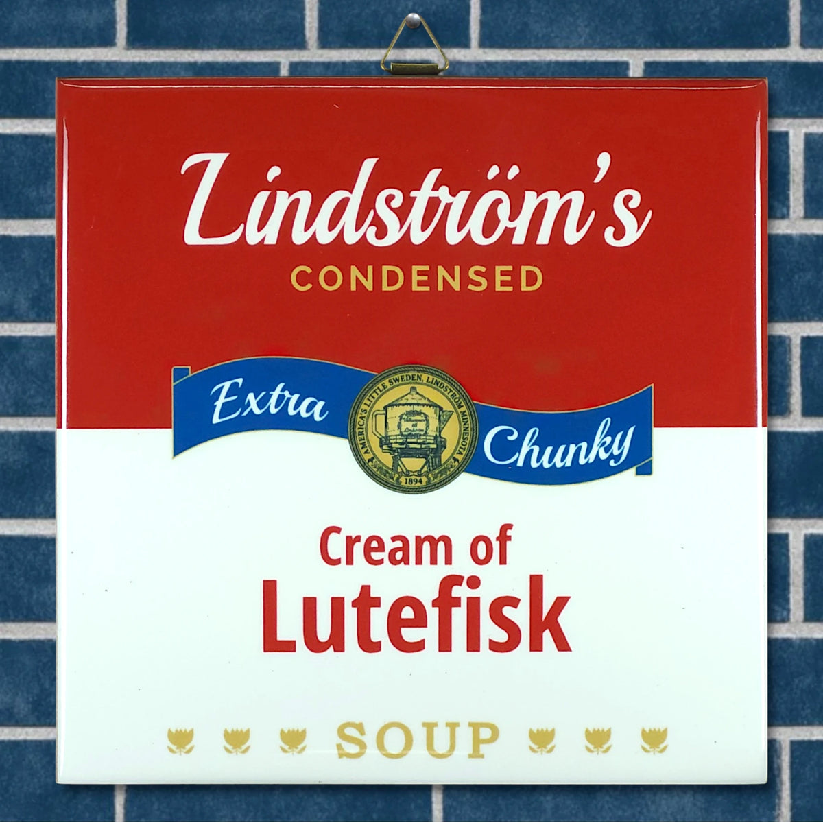 A can with a label reading Lindströms Condensed Extra Chunky Cream of Lutefisk Soup sits in front of white ceramic tile, featuring Minnesota folk art-inspired design elements.