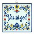 Cork-backed trivet with Norwegian and Swedish folk art patterns in blue, red, and yellow. The words “Var så god” are centered.