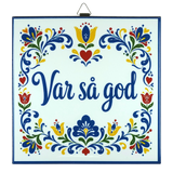 Cork-backed trivet with Norwegian and Swedish folk art patterns in blue, red, and yellow. The words “Var så god” are centered.