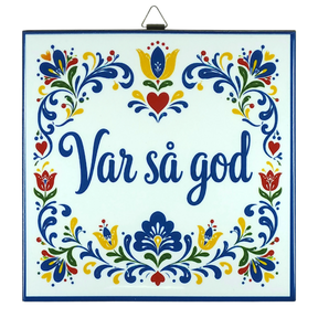 Cork-backed trivet with Norwegian and Swedish folk art patterns in blue, red, and yellow. The words “Var så god” are centered.