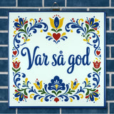 Ceramic trivet featuring blue floral patterns and the words “Var så god,” displayed on a blue tile background with Swedish folk art-inspired designs.