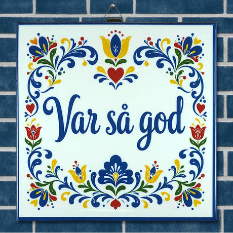 Ceramic trivet featuring blue floral patterns and the words “Var så god,” displayed on a blue tile background with Swedish folk art-inspired designs.