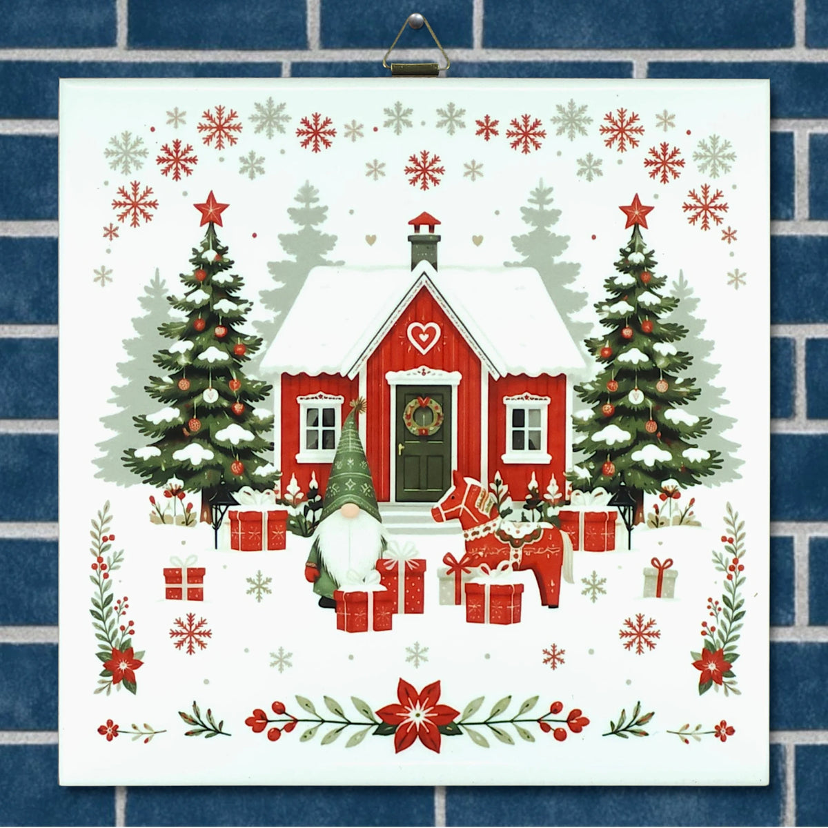 Scandinavian ceramic trivet with red house in snow, gnomes, gifts, trees, and snowflakes, surrounded by holiday greenery on a blue brick-patterned background.