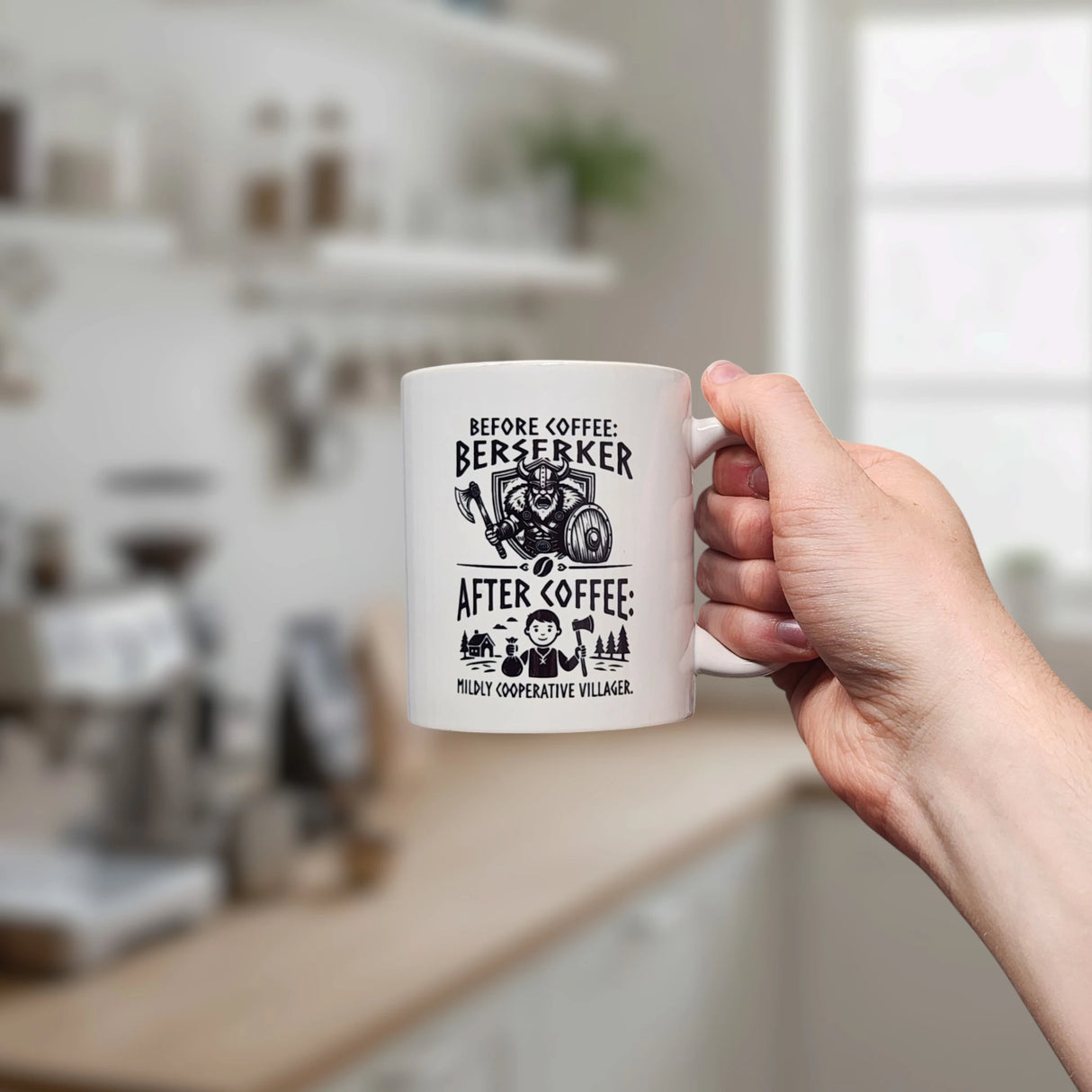 In a kitchen, a hand holds the Swedish Gift Store’s “Mug: Viking Berserker Before Coffee” 11 oz ceramic mug, featuring a wild Viking labeled "Before coffee" and a peaceful village scene labeled "After coffee.