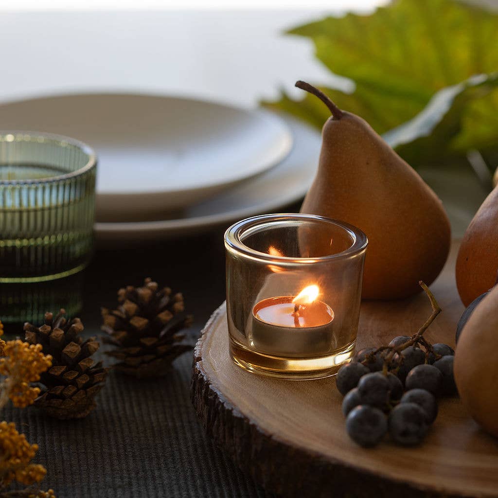 A lit Candle: Pure Beeswax Tea Light Candle – Handmade Raw / Clear Cup sits on a wooden slab, surrounded by pears, grapes, pinecones, and a green glass. Plates and autumn leaves in the background add to the cozy handmade ambiance.