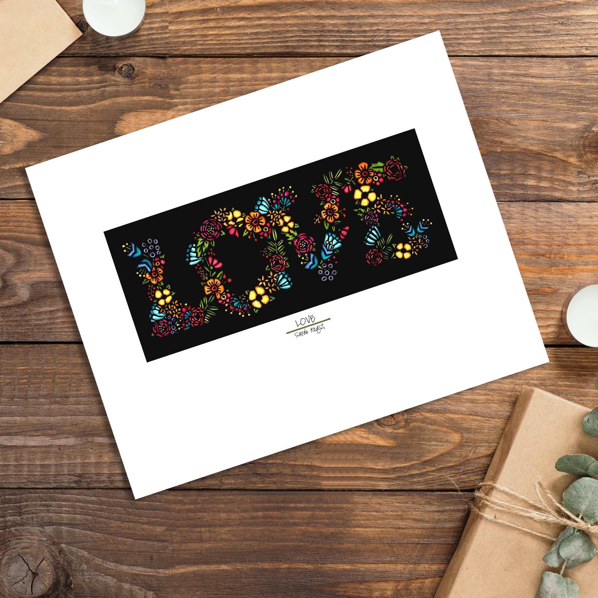 A charming scene with Sarah Angsts 8x10 LOVE print features a floral design on a black background, artfully placed on wood. Nearby, flickering candles and a wrapped gift enhance its elegance, making it perfect for any occasion.