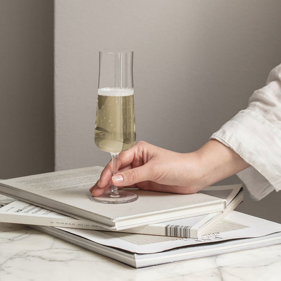 A hand holds a sleek Orrefors Informal Champagne Glass filled with sparkling wine, resting elegantly on an open book atop two closed ones on a marble surface, capturing the essence of Scandinavian design.