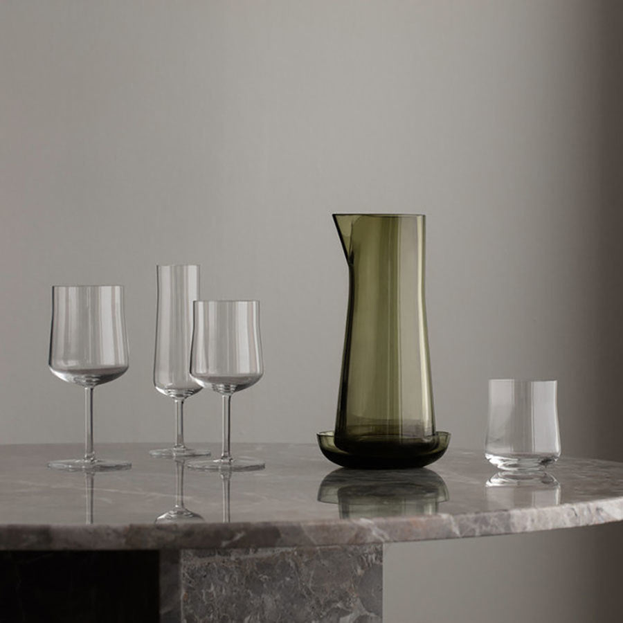 A green glass pitcher and four assorted glassware pieces, including an Orrefors Informal Champagne Glass from a 2-pack, are arranged on a marble surface against a neutral background, showcasing the simplicity and elegance of Scandinavian design.