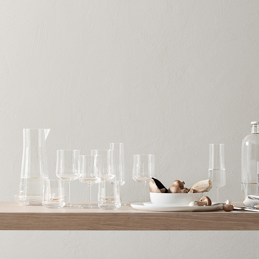 A wooden table displays assorted empty glasses, featuring the Orrefors Informal Champagne Glass (2-Pack), a carafe, a plate of mushrooms, and a bottle against a plain light wall—reflecting Scandinavian design simplicity.