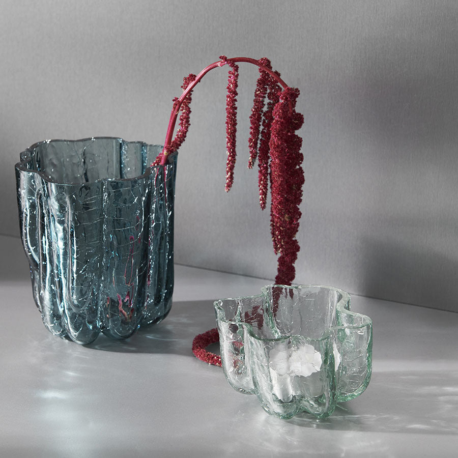 Two KostaBoda Crackle Bowl Votive Circular glass vases, evoking Åsa Jungnelius designs, sit elegantly on a table. One is serene blue, the other clear; a red flowering plant gracefully arches over the blue vase.