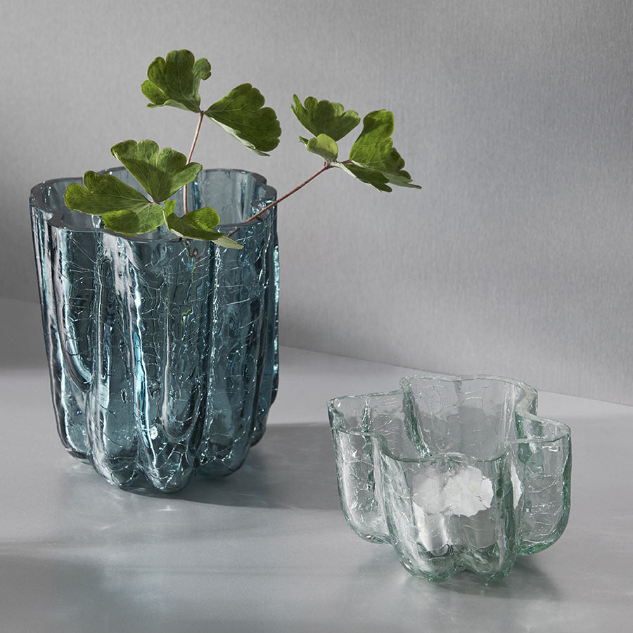 Two elegant KostaBoda Crackle Bowl Votives are on a surface; the left cradles green plant stems, and the right remains empty. Both feature a wavy, textured design reminiscent of sophisticated crystal glass artistry by Åsa Jungnelius.