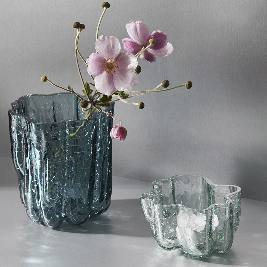 The KostaBoda: Crackle Bowl Votive Circular by Åsa Jungnelius is displayed elegantly on a smooth gray surface, showcasing its wavy crystal glass design. One bowl holds pink flowers on long stems, while the other remains empty.