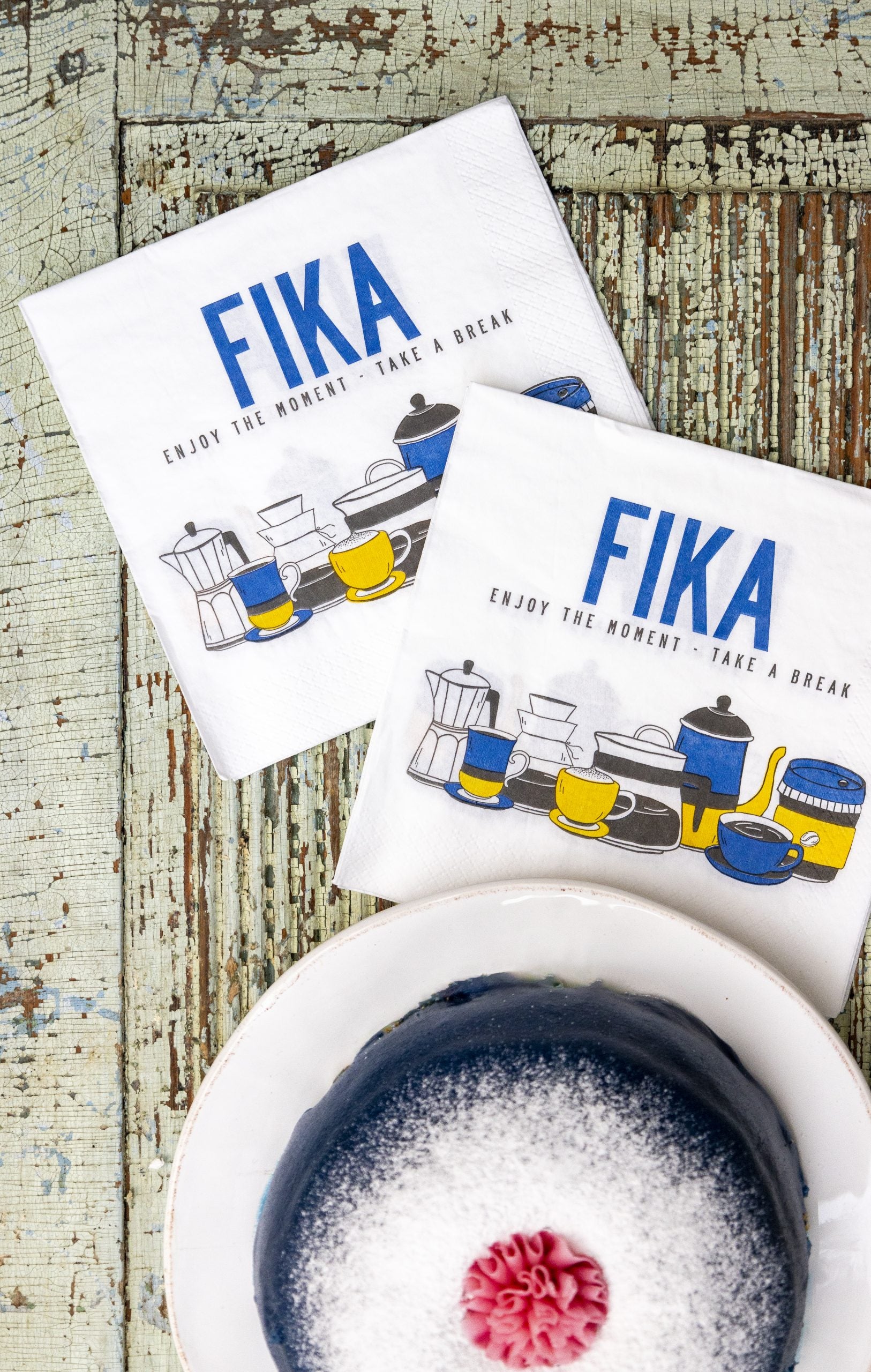 A small cake dusted with powdered sugar and topped with candy rests on a plate. Beside it, two Swedish Fika Retro Napkins from a 25-pack featuring vintage coffee pot and cup designs are placed on a rustic wooden surface.