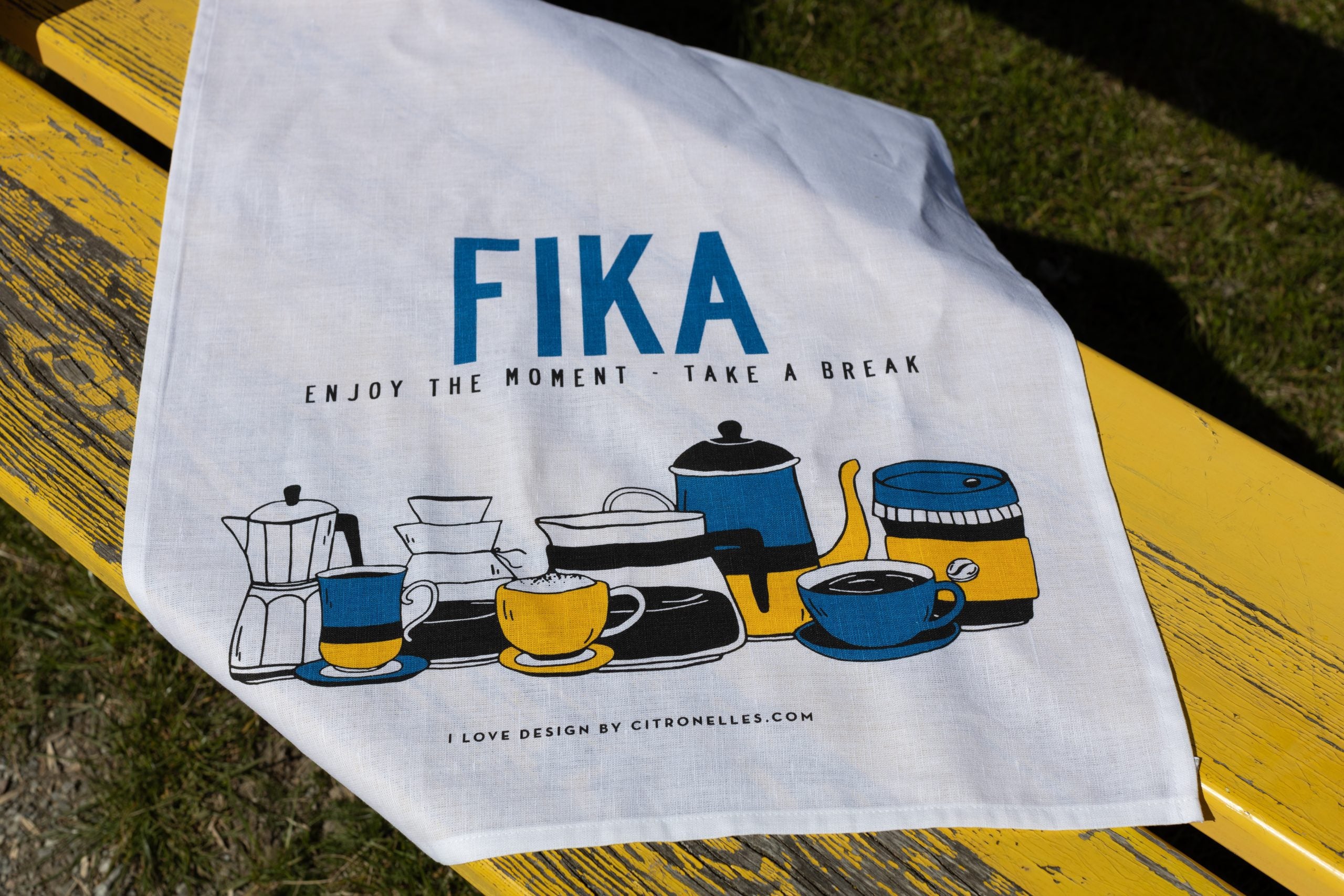 The Tea Towel: Fika Retro Tea Towel features retro coffee and tea illustrations on a yellow bench backdrop, with FIKA - Enjoy the Moment - Take a Break text. Its ideal for bringing cozy elegance to your kitchen décor.