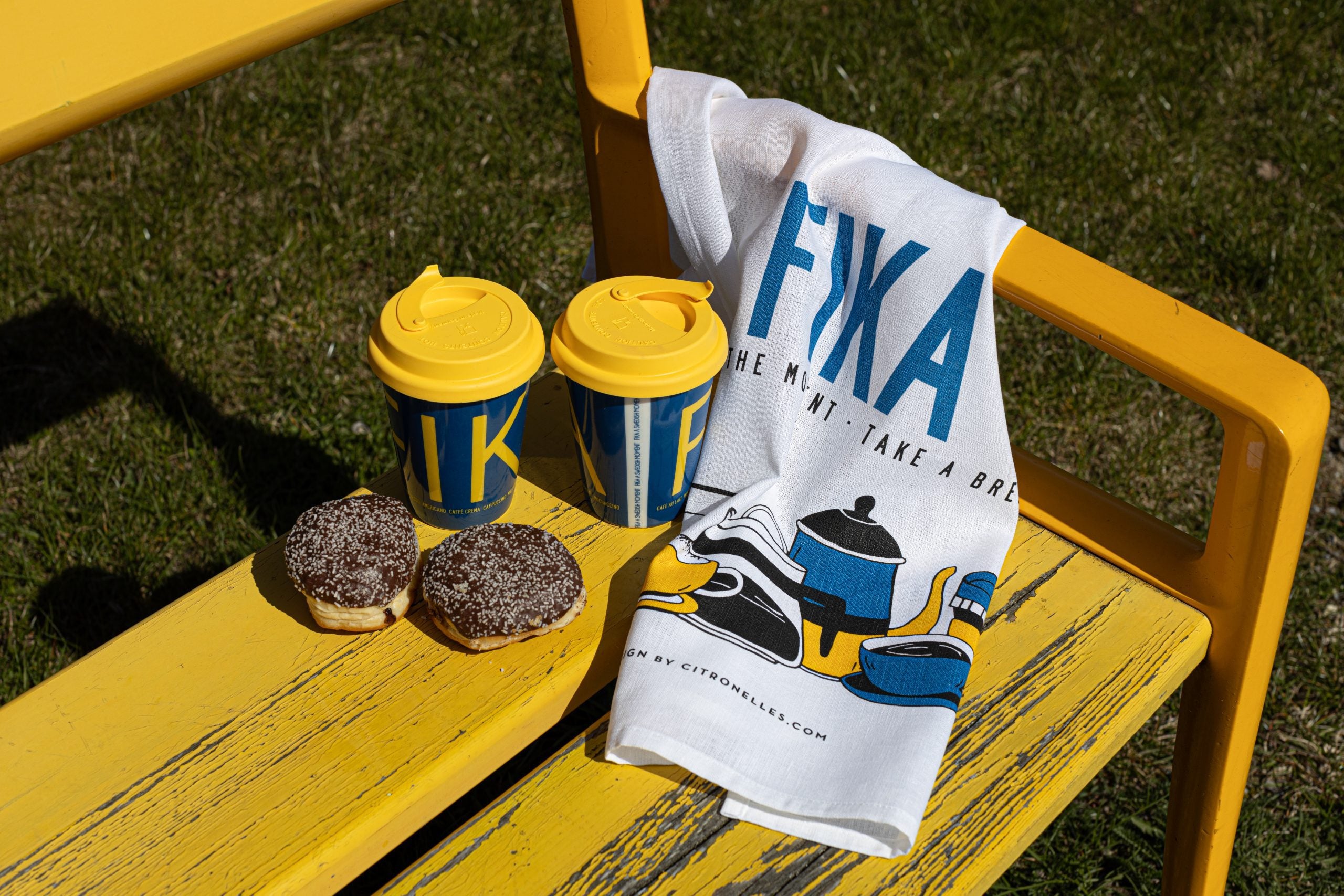 On a yellow bench, two yellow travel mugs and two chocolate donuts sit beside a white Swedish FIKA Retro Tea Towel with blue and yellow graphics, as swaying grass frames the charming scene.