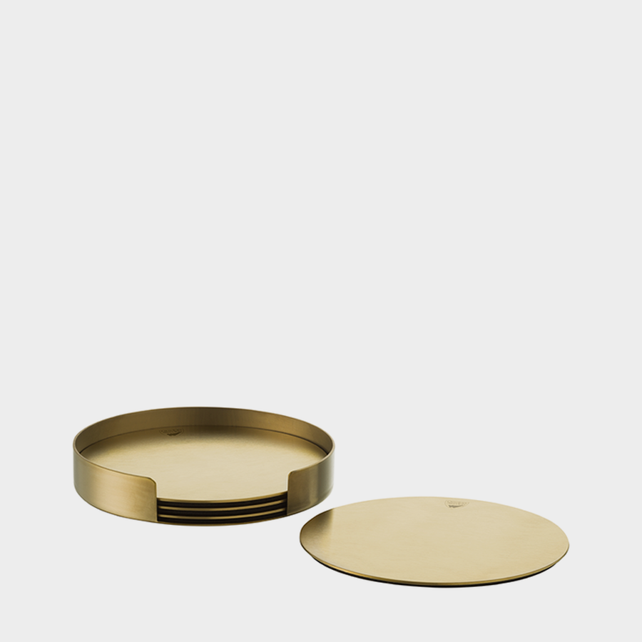 A set of Orrefors Peak Non Slip Coasters (4-Pack) in round, gold-colored metal, includes a sleek holder. One coaster sits beside the holder, reflecting Martti Rytkönens elegant style.