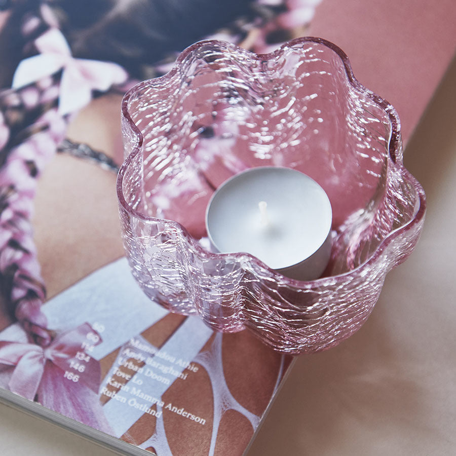 The KostaBoda: Crackle Bowl Votive Pink, reminiscent of Åsa Jungneliuss artistry, illuminates an open magazine page featuring a braided hairstyle and a chic white outfit.