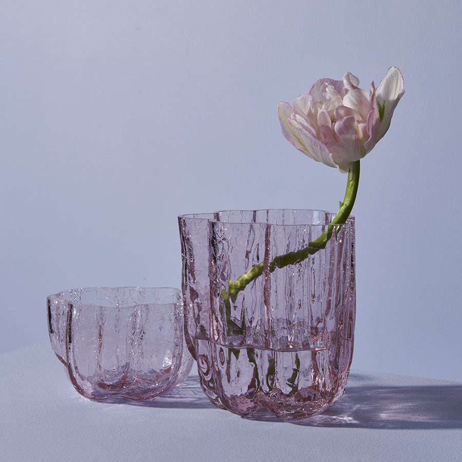 Pink glass vases, one holding a white and pink tulip, grace the scene alongside a KostaBoda Crackle Bowl Votive in Pink, designed by Åsa Jungnelius. The arrangement is serenely set against a plain backdrop.