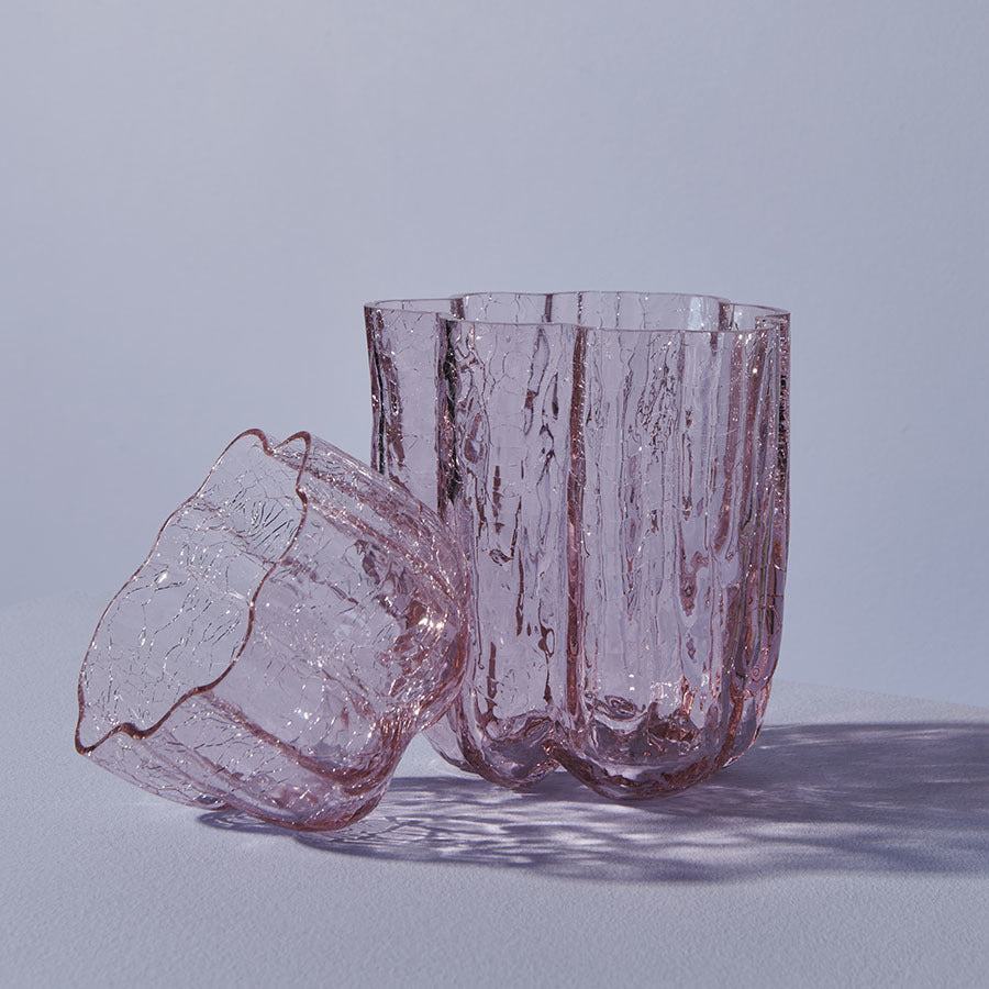 Two textured, transparent pink glasses, inspired by Åsa Jungnelius artistry, are displayed—one upright, the other on its side—evoking the elegance of a KostaBoda: Crackle Bowl Votive Pink. Their shadows dance gracefully on a light purple surface.