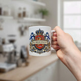 A hand displays the Nordic Wholesale Mug: Finland Coat of Arms Custom Name Ceramic Mug (11 oz Capacity), featuring a white finish with a detailed crest, two Finnish flags, and "Korhonen" above "Suomi" on the front. Set against a bright, contemporary kitchen backdrop with blurred shelves and a window, this mug is an ideal personalized family gift for anyone proud of their Finnish heritage.