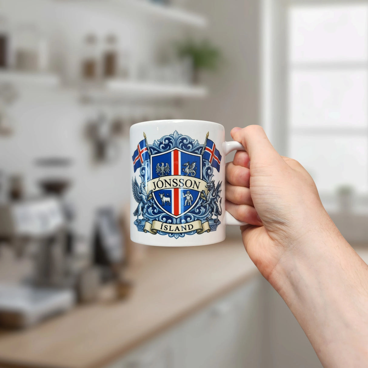 A hand holds the Nordic Wholesale Mug: Iceland Coat of Arms Custom Name Ceramic Mug (11 oz Capacity), featuring a white design personalized with the name Jonsson, an ornate blue crest, two Icelandic flags, and the word "Island." A blurred kitchen appears in the background.