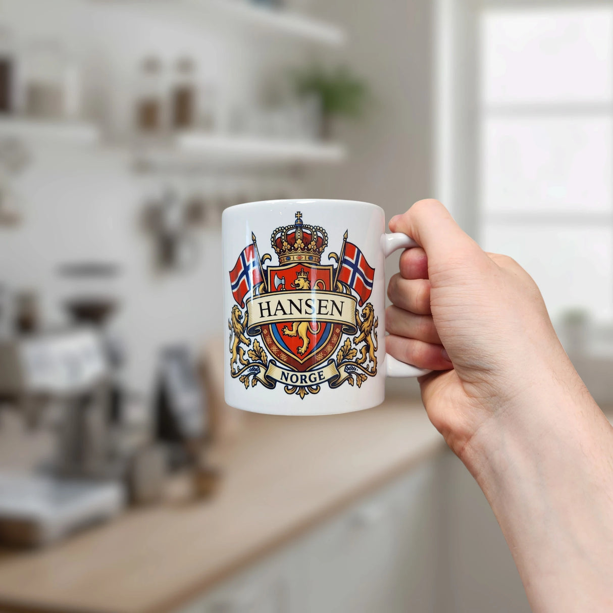 A hand holds the Mug: Norway Coat of Arms Custom Name Ceramic Mug (11 oz Capacity) by Nordic Wholesale, personalized with the Norwegian coat of arms, two Norwegian flags, and the words “Hansen” and “Norge.” A blurred kitchen with shelves and a window forms a cozy backdrop, highlighting this unique Norway heritage gift.