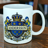 The Mug: Sweden Coat of Arms Custom Name Ceramic Mug (11 oz Capacity) by Nordic Wholesale is a white ceramic mug featuring a blue and gold crest with the name Andersson, Swedish flags, lions, and a crown. The word "Sverige" is written below. This personalized Sweden mug is displayed on a wooden table.