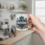 A hand holds the Mug: Sweden Coat of Arms Custom Name Ceramic Mug by Nordic Wholesale—an 11 oz white ceramic mug featuring a royal blue and gold crest, Swedish flags, and a customizable name (shown here as Andersson above Sverige)—set against a bright, blurred kitchen background.