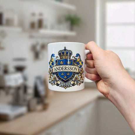A hand holds the Mug: Sweden Coat of Arms Custom Name Ceramic Mug by Nordic Wholesale—an 11 oz white ceramic mug featuring a royal blue and gold crest, Swedish flags, and a customizable name (shown here as Andersson above Sverige)—set against a bright, blurred kitchen background.