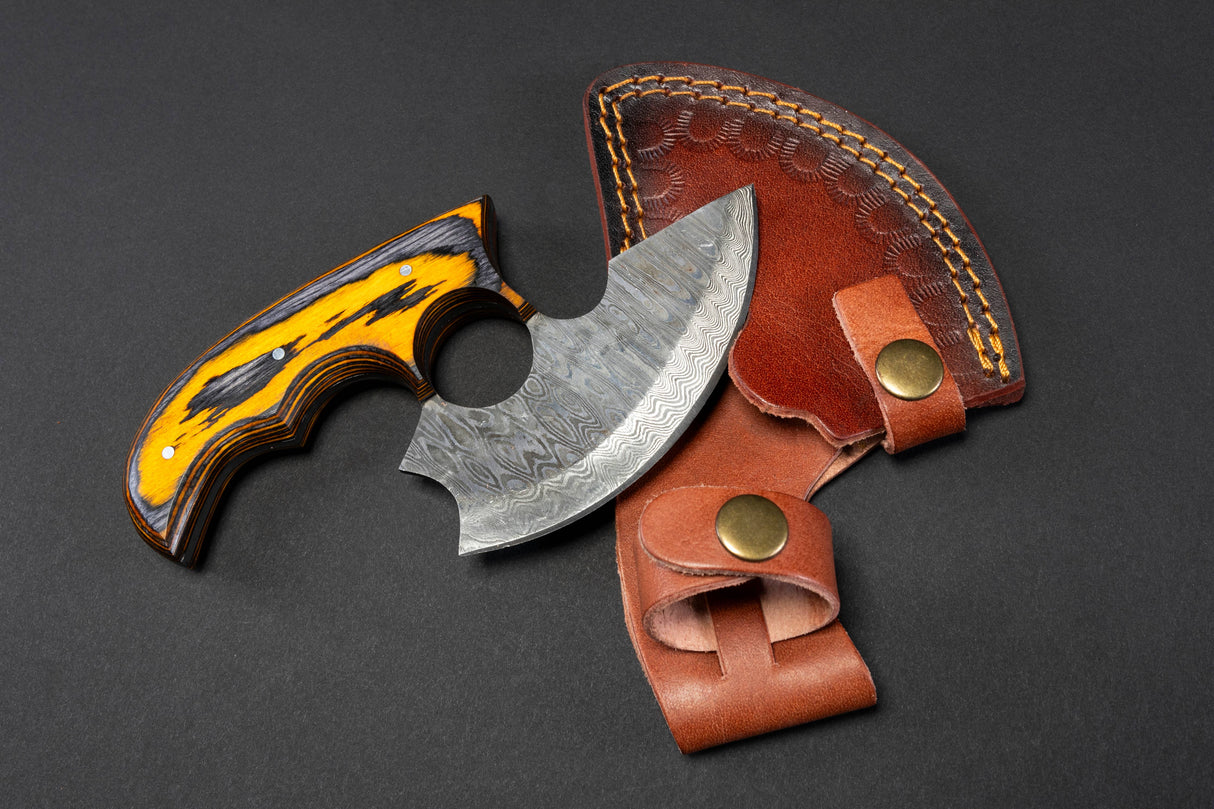 The Alaskan Ulu Style Blade, featuring a patterned High Carbon Damascus Steel curved blade and an ergonomic yellow-black handle, is shown beside its brown leather sheath on a dark surface.