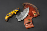 The Alaskan Ulu Style Blade, featuring a patterned High Carbon Damascus Steel curved blade and an ergonomic yellow-black handle, is shown beside its brown leather sheath on a dark surface.