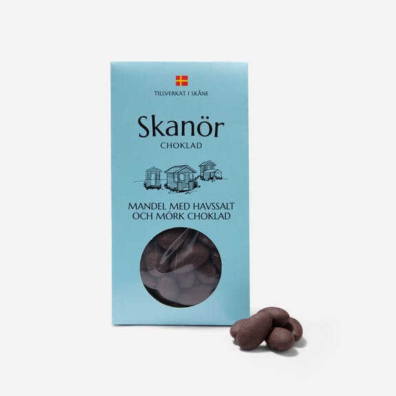 A box of Candy: Skanör - Dark Chocolate Almonds with Sea Salt (100g) features a blue design, highlighting the blend of almonds and sea salt wrapped in dark chocolate. Three chocolate-covered almonds are displayed gracefully in front of the box.