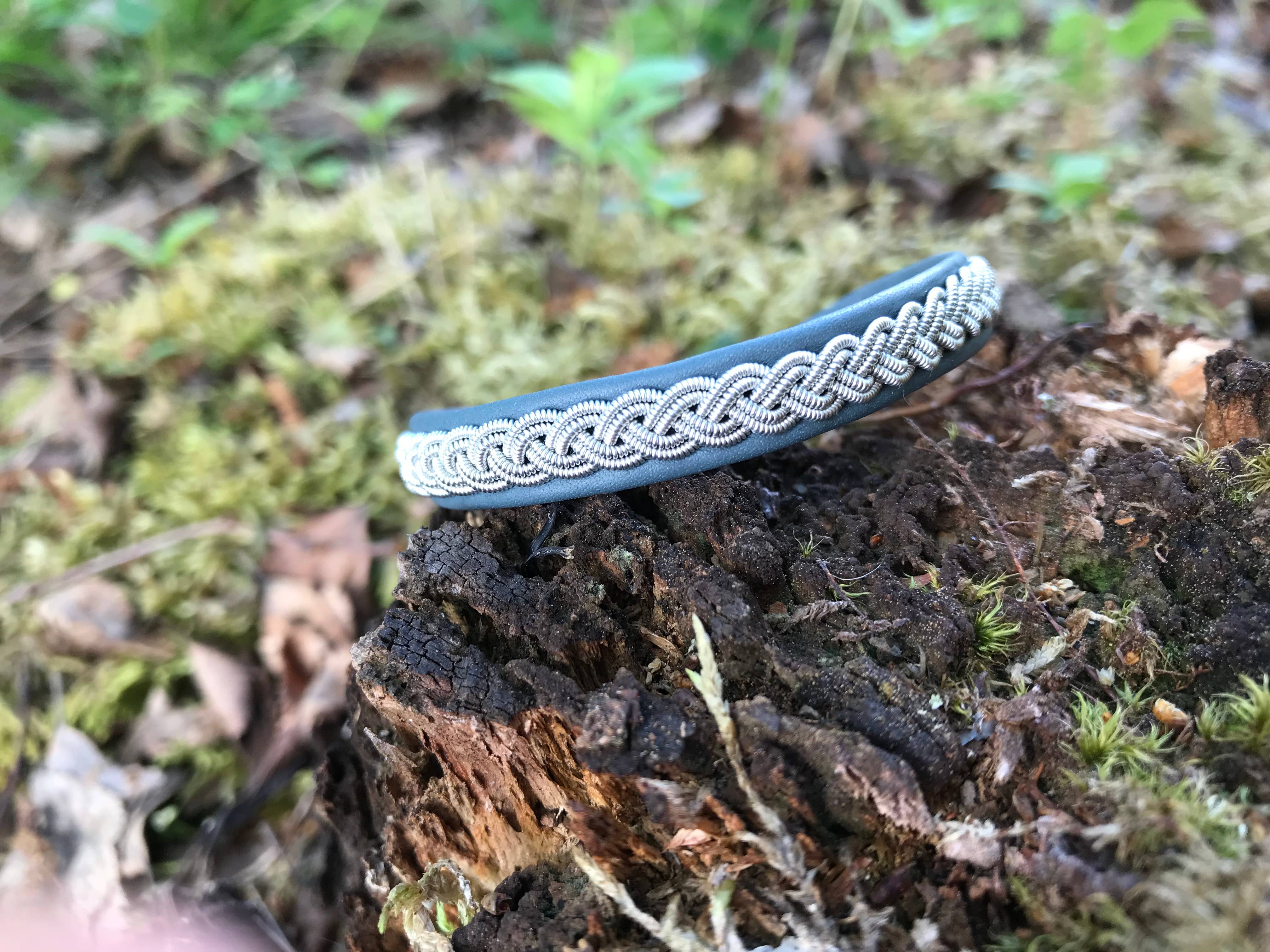 The Bracelet: Lapland Sami Tin Bracelet Single Braid #1: Blue Gray, crafted in traditional style, rests on a mossy tree stump outdoors with green foliage in the background.