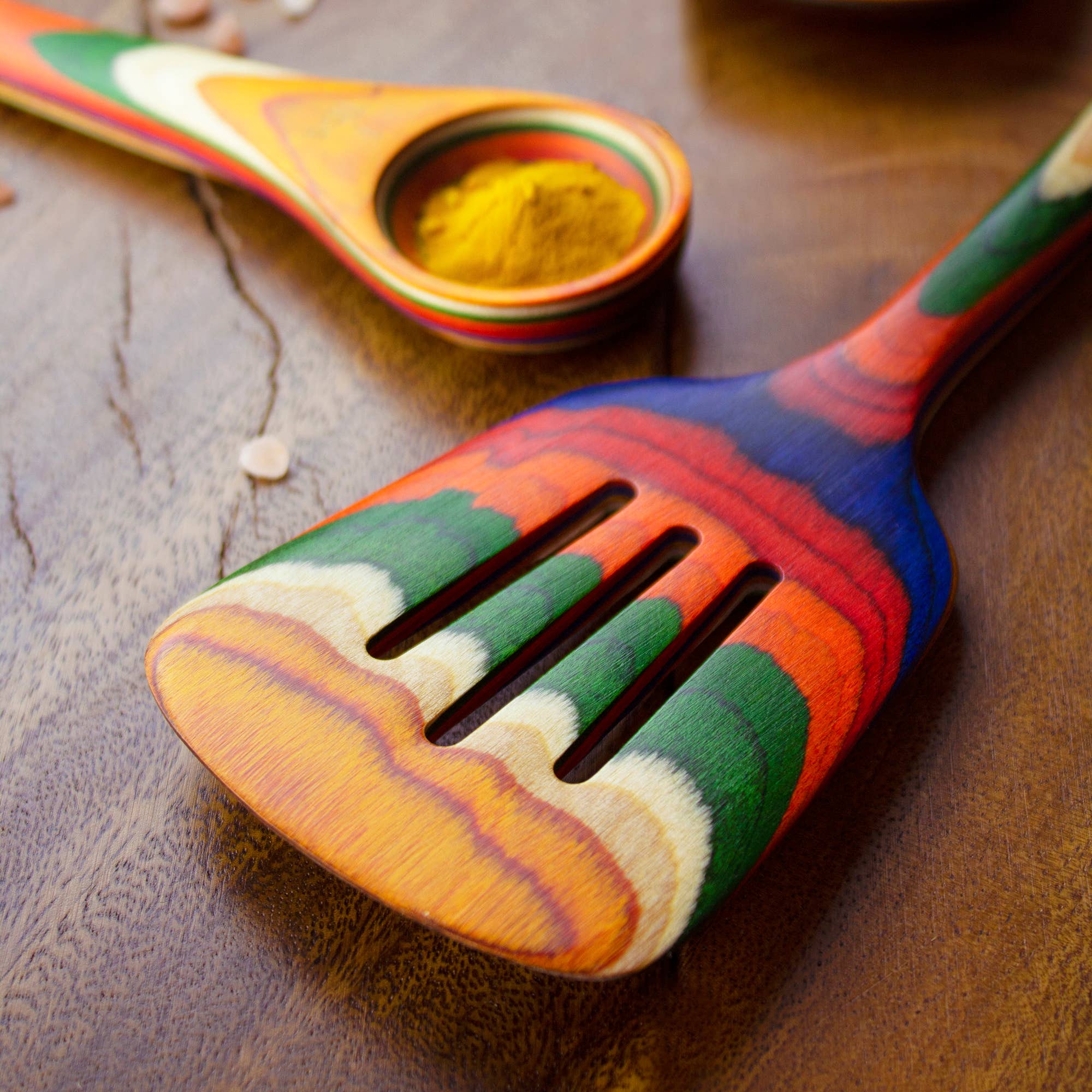 A lively slotted spatula from the Utensil: Marrakesh Collection rests on a wooden surface, accompanied by a colorful measuring spoon containing a hint of turmeric.