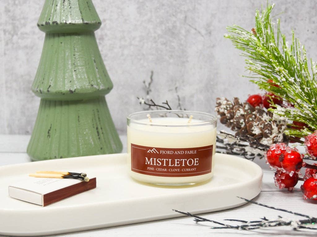 A Mistletoe Premium Soy Candle (4oz, 2 wicks, 20+ hour burn) by Fjord & Fable sits on a tray with matches; a green tree ornament and holiday greenery with red berries complete this festive Christmas candle scene.